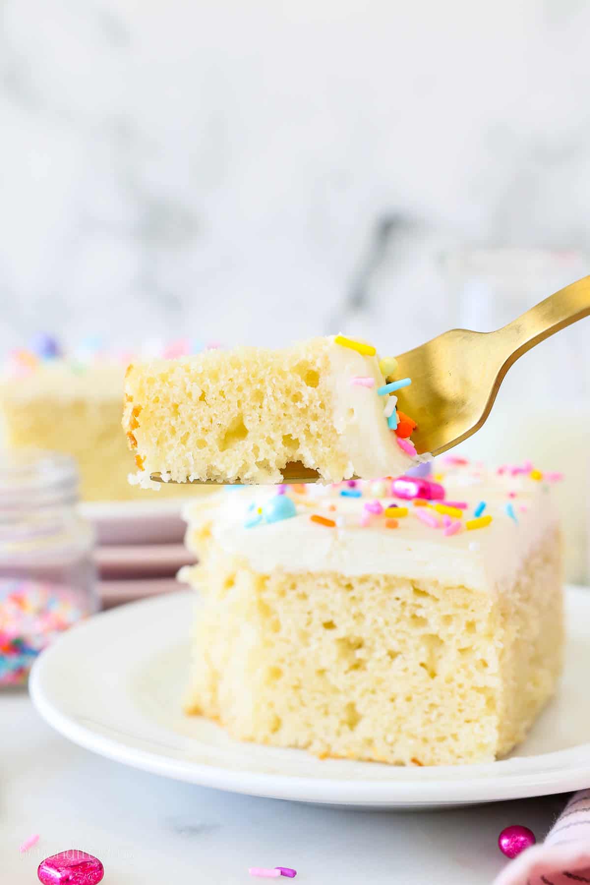 This moist vanilla cake recipe is perfectly sweet and fluffy and covered with creamy vanilla frosting. It's the best cake for any occasion! A forkful of vanilla cake held up in front of a slice of frosted cake garnished with rainbow sprinkles.