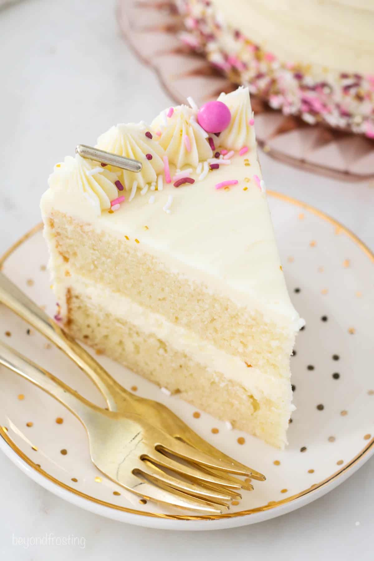 Learn how to make a layer cake with this easy step-by-step tutorial. Everything you need to know, from leveling cakes to frosting the layers! A slice of frosted layer cake decorated with frosting swirls and rainbow sprinkles on a white plate next to a fork.