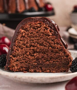 A slice of glazed chocolate pound cake on a plate next to cherries and blackberries, with the rest of the pound cake on a cake stand in the background.