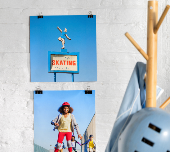 Two photo prints hanging on a white wall, featuring a skating sign and a girl roller skating. A hat rack to the side of the photos has a helmet and shirt hanging from it.