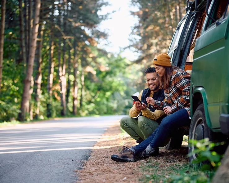 Two people looking at their device sitting out the door of their van, on the side of the road in a forested area.