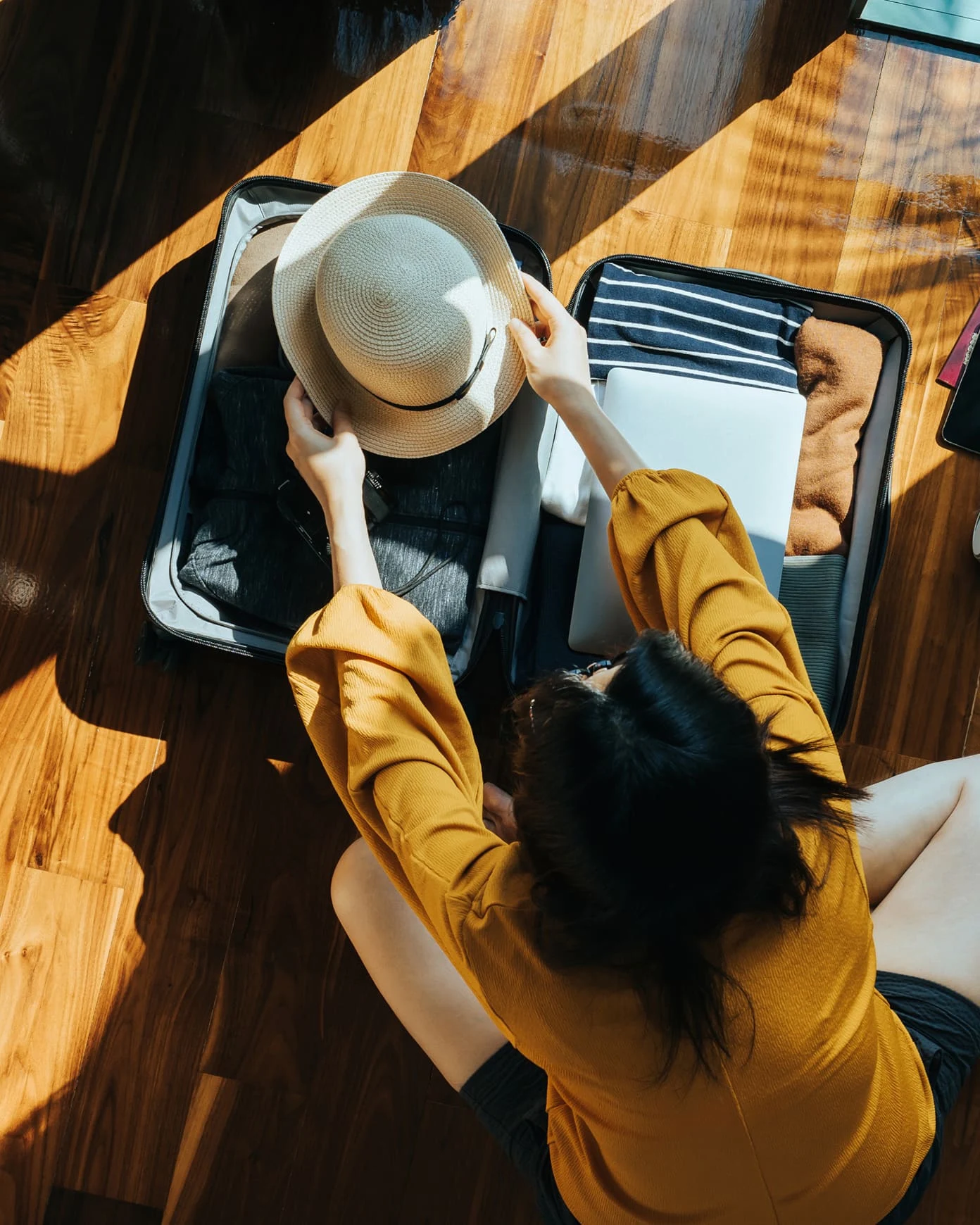 An aerial view of an individual putting a beige sun hat in their suitcase.