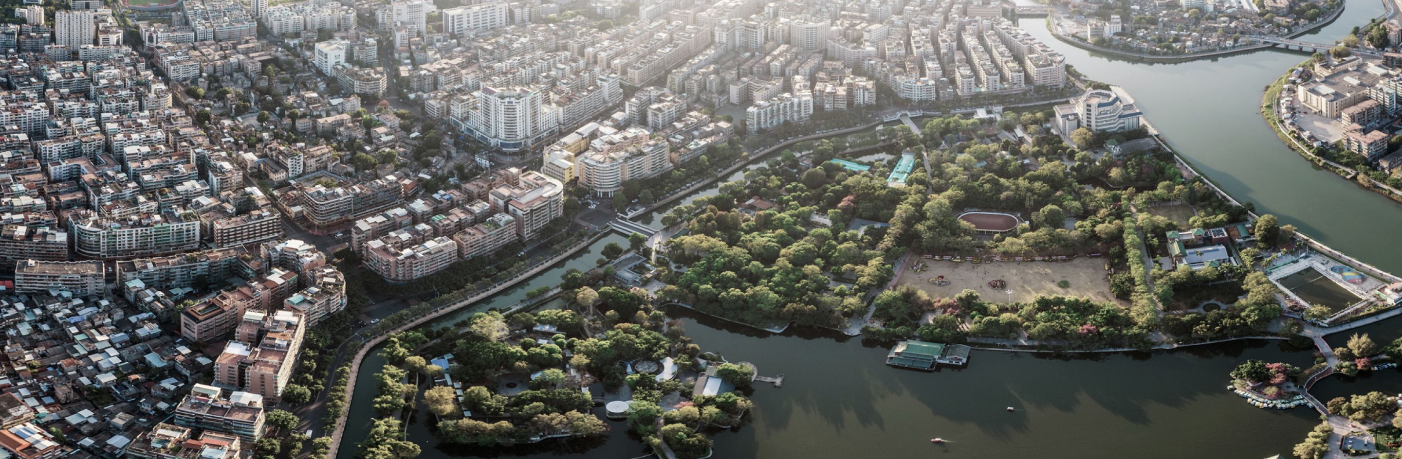 Bird's-eye view of a dense city bordering a large, lush park and a winding river under soft sunlight.