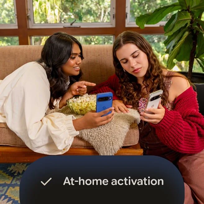 Two women sitting on a couch with popcorn, looking at a Google Pixel 10 in Indigo, and a Google Pixel 10 Pro in Porcelain, with an overlay reading "At-home activation" with a checkmark icon.