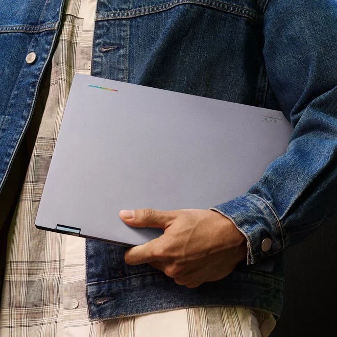 A person in a denim jacket holding a closed Acer laptop under their arm.