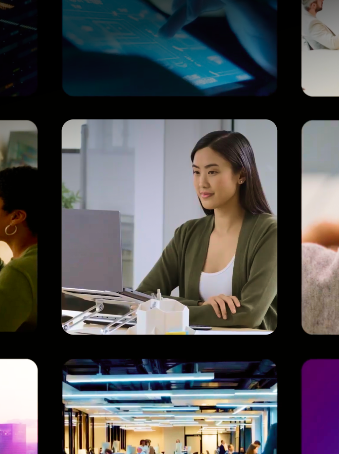 A young professional woman smiles while working on a laptop at her desk in a bright, modern office.