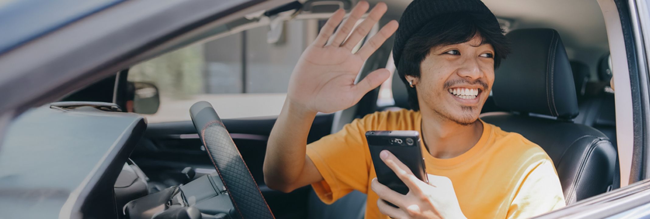 A smiling man in a yellow shirt and black beanie waving from a car's driver seat while holding a smartphone.