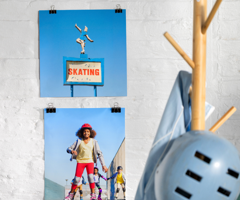 Two photo prints hanging on a white wall, featuring a skating sign and a girl roller skating. A hat rack to the side of the photos has a helmet and shirt hanging from it.