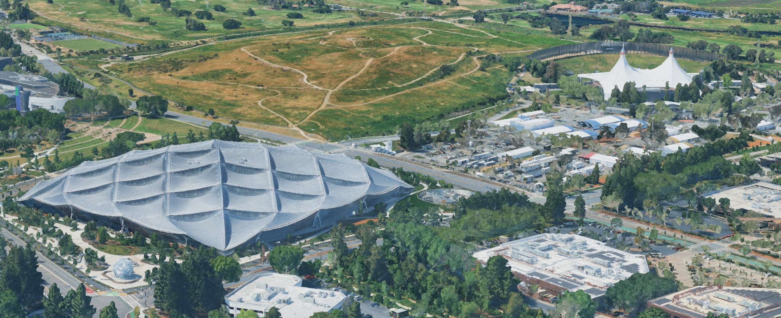 Aerial view: Google's Bay View campus with distinctive gray roof, Shoreline Amphitheatre, green fields, and roads.