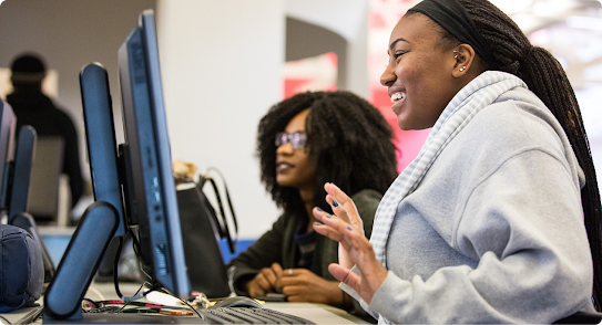 Two students work together at computers in a classroom, one smiling and speaking, suggesting a collaborative or learning environment.