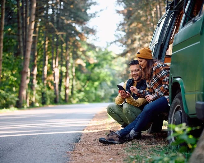 Two people looking at their device sitting out the door of their van, on the side of the road in a forested area.