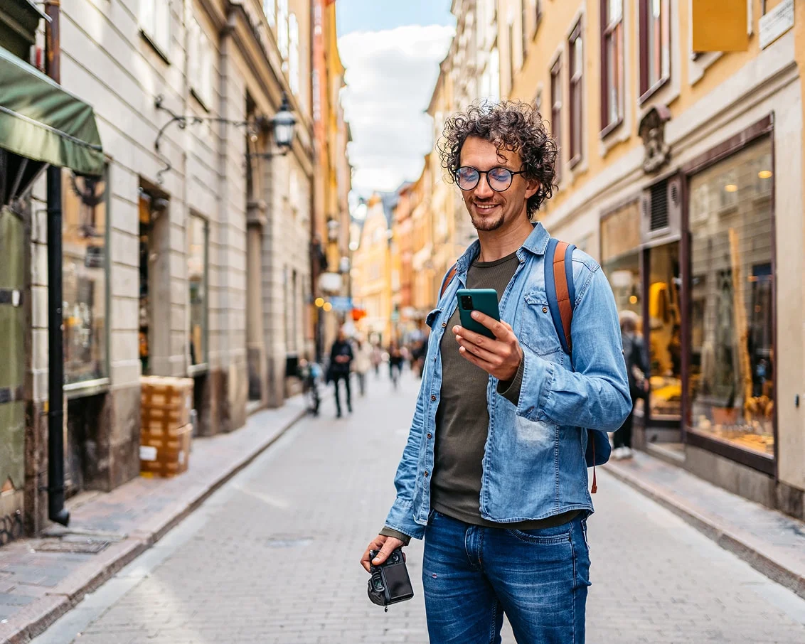 An individual standing on the street looking at his device and holding a camera.