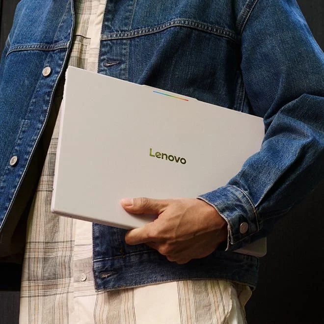 A person in a denim jacket holding a closed Lenovo laptop under their arm.