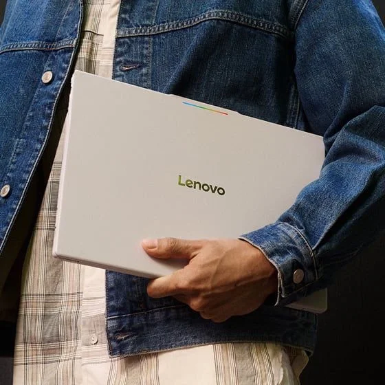 A person in a denim jacket holding a closed Lenovo laptop under their arm.