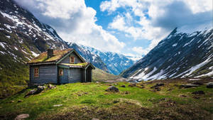En liten trehytte plassert i en fjell dal med snødekte topper og grønt gress. — 3840x2160 Bakgrunner