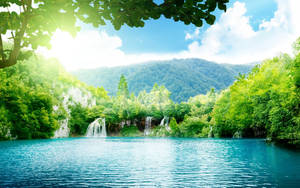 Turquoise lake with waterfalls surrounded by green forest and mountains under bright sun.