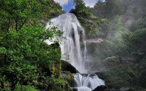 A majestic waterfall flows through a dense green forest with mist rising around rocks.