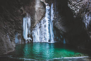 Frozen waterfall flows into a clear turquoise pool in a rocky canyon.