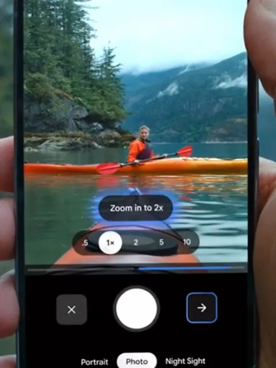 A hand holds a Pixel phone with the camera app open, showing a kayaker on a lake. The zoom function is being used to "Zoom in to 2x."