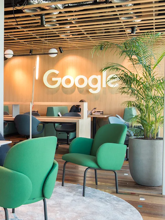 Zwei Personen sitzen in einem Arbeitsbereich – im Hintergrund ist das Google-Logo zu sehen.
