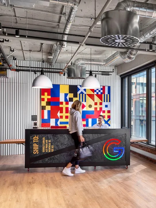 Eine Person geht an einer Rezeption in einer Bürolobby mit einem farbenfrohen, flaggenartigen Kunstwerk vorbei. Rechts davon steht in großen roten und orangen Neonbuchstaben „Google Montréal“.