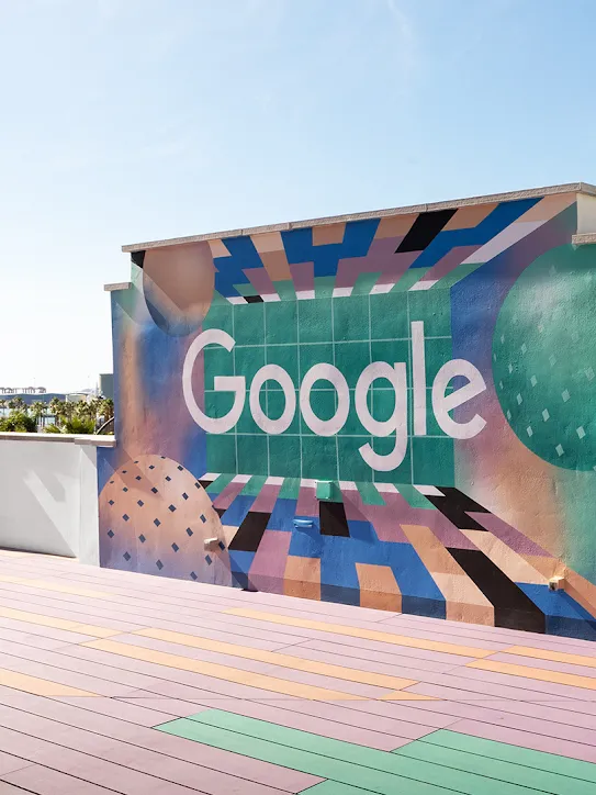 Eine Dachterrasse mit einer Wand, auf der das Google-Logo angemalt wurde. Im Hintergrund sind Palmen und Boote zu sehen.