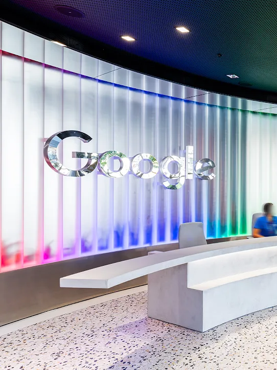 Eine geschwungene Wand mit silbernem Google-Logo wird rot, blau und grün beleuchtet. Davor sitzt eine Person hinter einer weißen Empfangstheke, an der gerade drei Personen vorbeigehen.